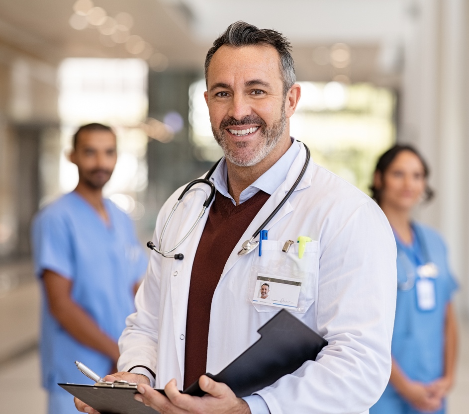 En el primer plano se ve un médico sonriente, en el segundo plano se ven un hombre y una mujer profesionales de la salud que también sonríen. Todos se encuentran en un pasillo de una clínica. En el primer plano se ve un médico sonriente, en el segundo plano se ven un hombre y una mujer profesionales de la salud que también sonríen. Todos se encuentran en un pasillo de una clínica.