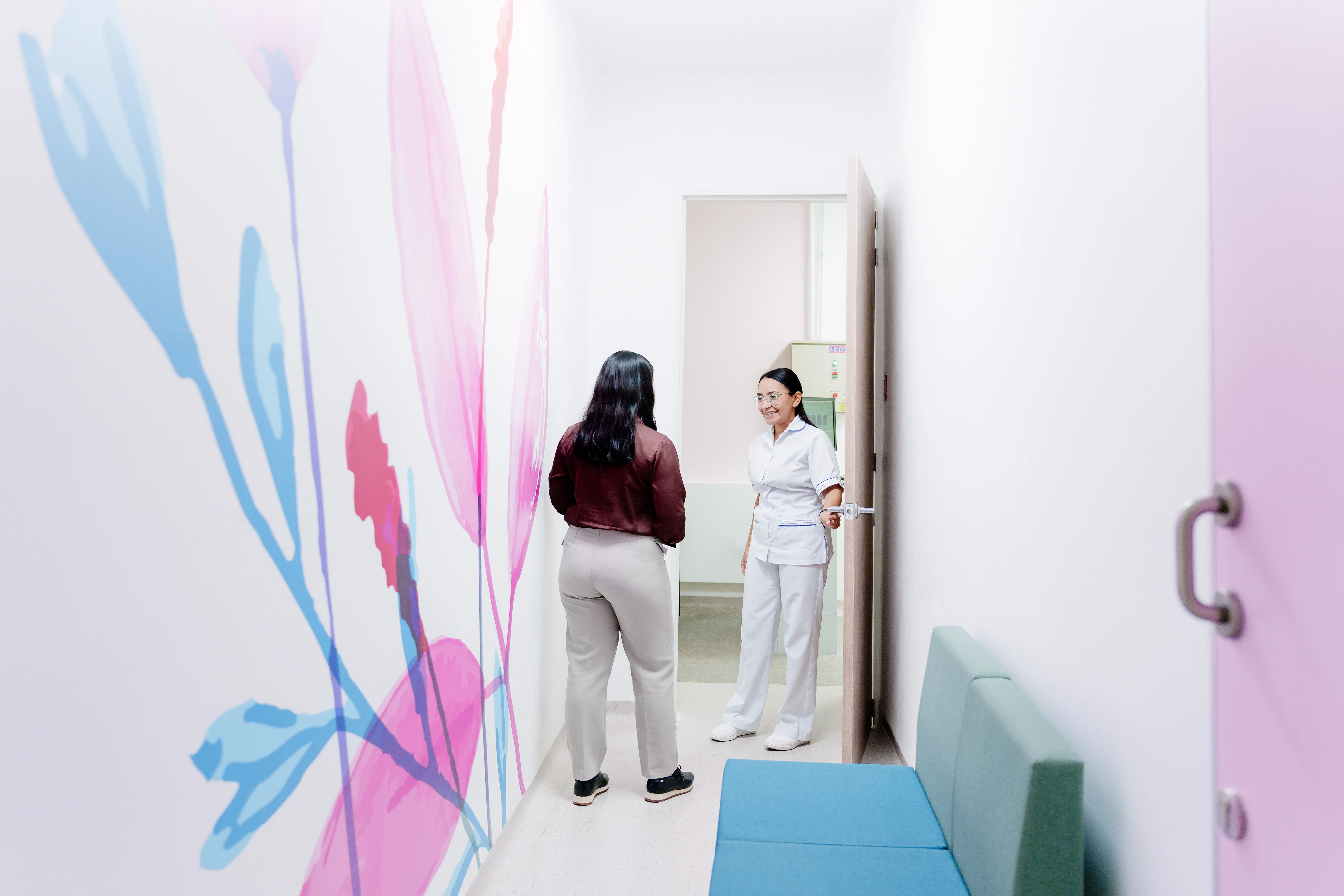 Hay un pasillo que en su pared izquierda tiene un mural de flores, al final hay una puerta en la cual una profesional de la salud le dala bienvenida a una paciente. Hay un pasillo que en su pared izquierda tiene un mural de flores, al final hay una puerta en la cual una profesional de la salud le dala bienvenida a una paciente.